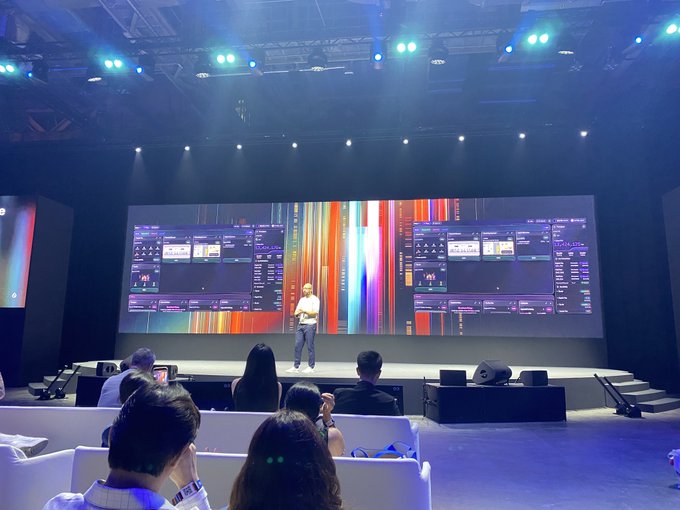 A speaker presents on stage at a tech event with large LED screens displaying colorful data visualizations and trading interfaces behind him, while attendees watch from seating in the foreground.
