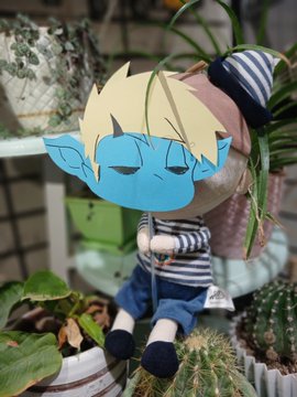 A plush toy of a blue-skinned character with blonde hair and pointed ears sits among potted plants and cacti on a shelf.