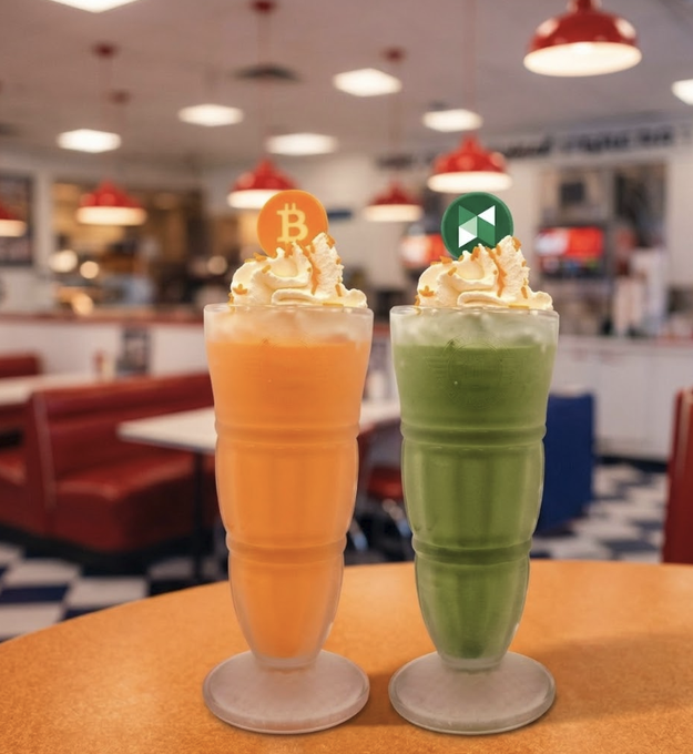 Two tall milkshakes in diner-style glasses topped with whipped cream, one orange with a Bitcoin 'B' symbol topper and one green with an Ethereum topper.