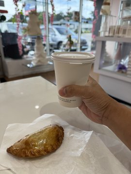 A hand holds a white paper coffee cup with a brown sleeve in what appears to be a café, with a glazed pastry on white wrapping paper in the foreground.