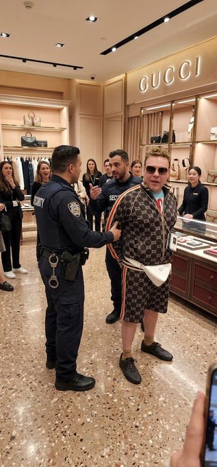A man in a Gucci outfit and sunglasses stands inside a Gucci store while a police officer in uniform holds his arm, with onlookers and staff watching the scene.
