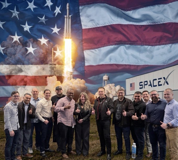 A group of about a dozen people pose giving thumbs-up in front of a SpaceX facility with a large American flag and rocket launch backdrop. The image relates to the post's patriotic theme about protecting Elon Musk's identity, featuring what appears to be a SpaceX team gathering at their launch site.