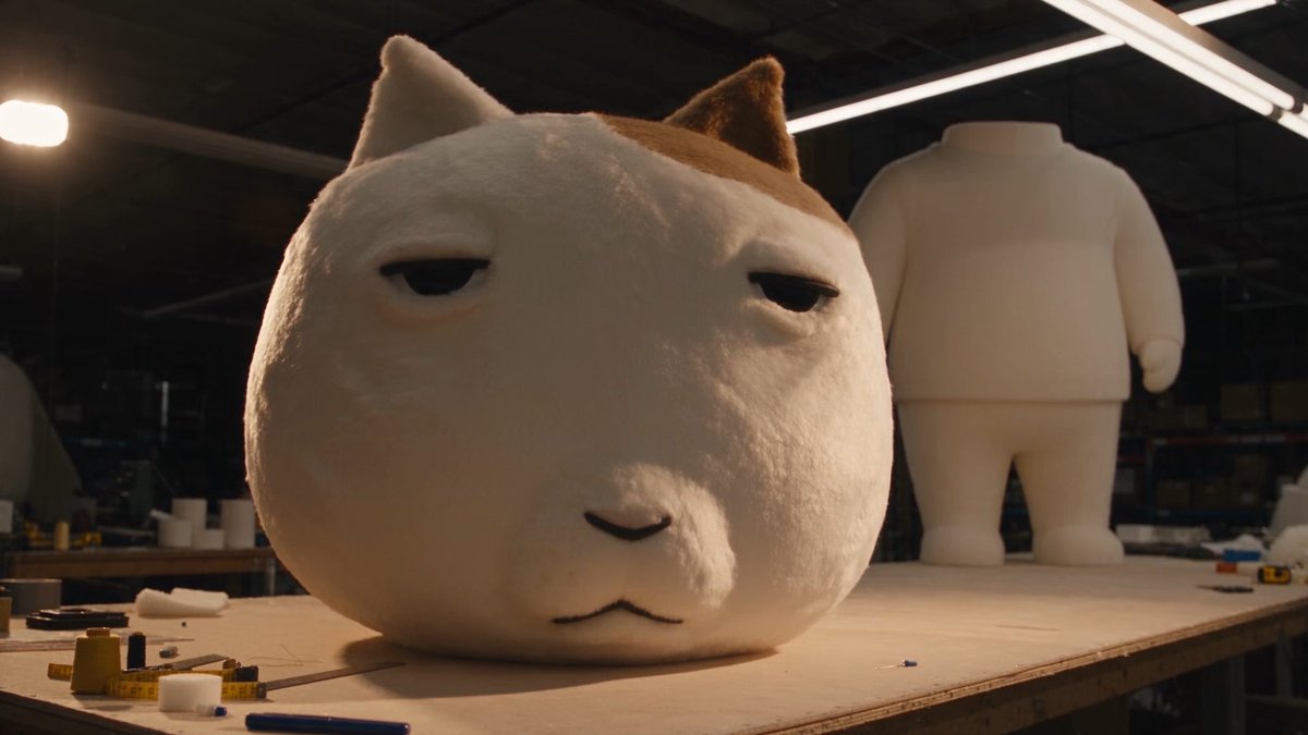 A detailed, plush cat head sculpture with an unimpressed expression sits on a workbench next to an unfinished white figure, surrounded by crafting tools and supplies. The image evokes the post's message, showcasing tangible, handcrafted artistry in response to skeptics claiming AI involvement.