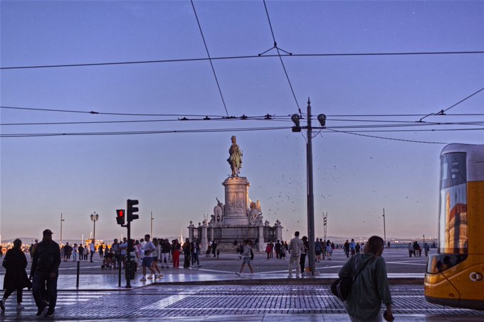 A dusky evening scene of a large public square featuring an equestrian statue, with pedestrians, tram wires, and a yellow tram visible.