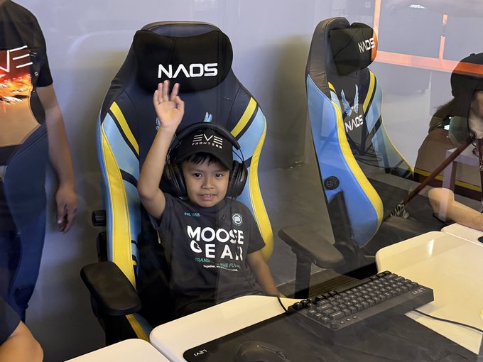 A young boy wearing a 'MOOSE GEAR' shirt and a gaming headset waving his hand while sitting in a NAOS-branded gaming chair in an esports setup.