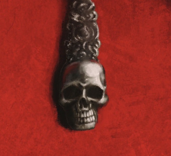 A dark metallic skull pendant with a decorative crown-like top rests against a deep red velvet background.