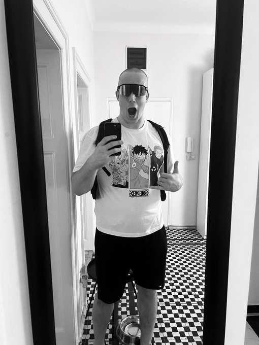 A black-and-white mirror selfie of a man wearing sunglasses, an anime graphic t-shirt, and a backpack, with his mouth open in an excited expression.