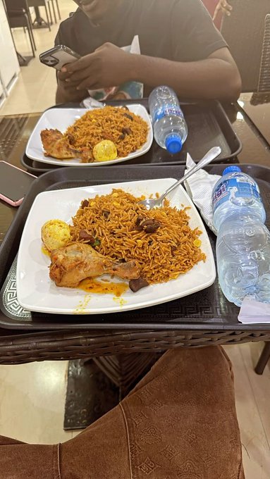 Two trays of food sit on a restaurant table, each containing a plate of orange-colored rice, a boiled egg, and chicken, accompanied by bottled water, while a person in a green shirt uses a phone in the background.