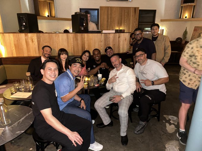 Group photo of approximately ten smiling people posing together at an indoor venue, with a man in the foreground giving a peace sign and a white birthday cake visible on the table.