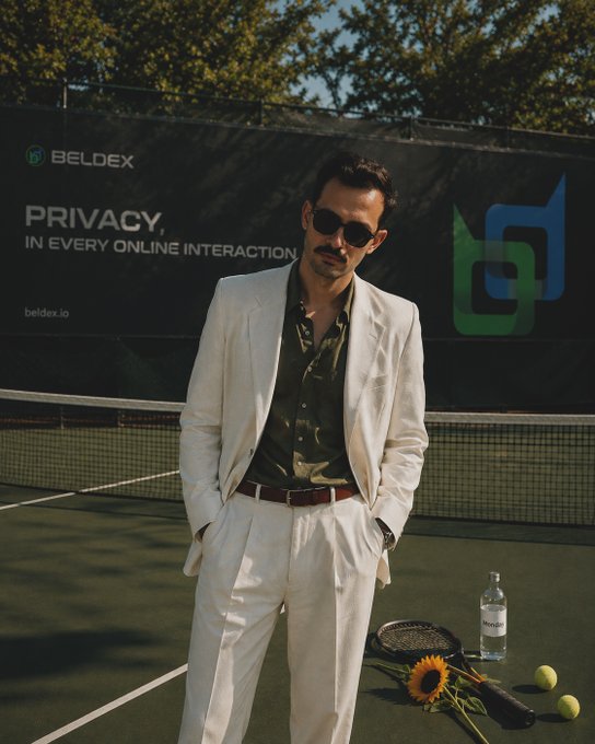 A man in a cream suit and sunglasses stands on a tennis court beside a racket, sunflower, water bottle, and tennis balls, with a "BELDEX - Privacy in Every Online Interaction (beldex.io)" banner displayed behind him. The image visually reinforces the post's message about Beldex's layered, multi-quorum network architecture being a coordinated team effort, much like the composed, multi-element scene around the figure.