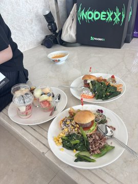 A meal of sandwiches, a small burger, salad, and side dishes is laid out on a table next to a bag with the text 'NODEXX'.