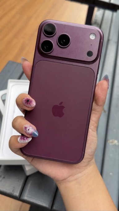 A hand with floral manicured nails holds up a purple iPhone with its camera module and Apple logo visible on the back.