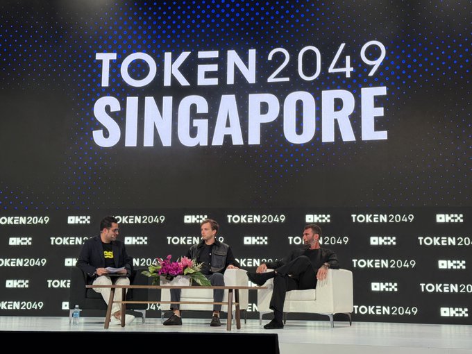 Three men sit in chairs on stage at the TOKEN2049 Singapore conference, engaged in a panel discussion against a backdrop displaying "TOKEN2049 SINGAPORE" alongside OKX logos, consistent with a post referencing the @token2049 event.
