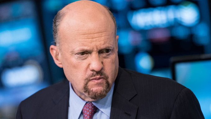 A close-up of a bald man with a goatee wearing a dark suit, light blue shirt, and patterned tie, appearing serious in what looks like a news studio with blurred monitors in the background.