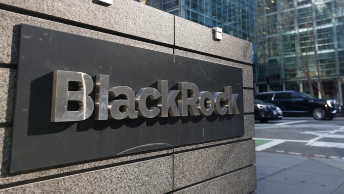 A polished stone sign displays the metallic "BlackRock" logo outside the company's corporate building, with a black SUV and modern glass skyscrapers visible in the background. The image visually represents BlackRock's physical corporate presence as context for the announcement of its new partnership with Ondo Finance to launch tokenized investment portfolios.