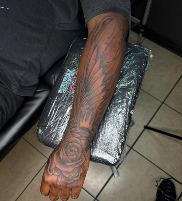A person's tattooed arm featuring a large rose and wing design rests on a plastic-wrapped tattoo chair in a studio.