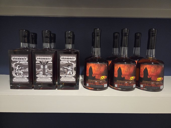 A collection of Smolrun whiskey bottles sits on a white shelf against a dark blue wall, with three "Standoff" bottles on the left featuring black and white illustrated labels and three "Styx" bottles on the right with fiery orange and black artwork depicting a hooded figure, matching the post's discussion comparing the two varieties.