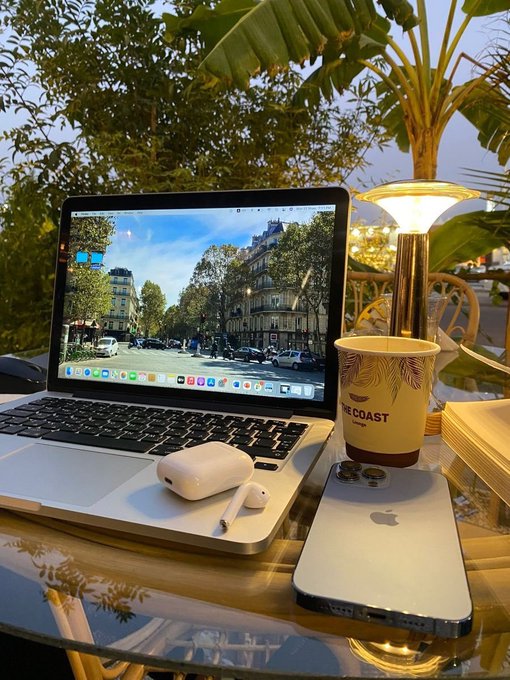 A MacBook laptop with AirPods and an Apple Pencil sits on a glass table next to an iPhone and a coffee cup labeled "THE COAST," displaying a Parisian street scene wallpaper with a lamp and plants in the background.