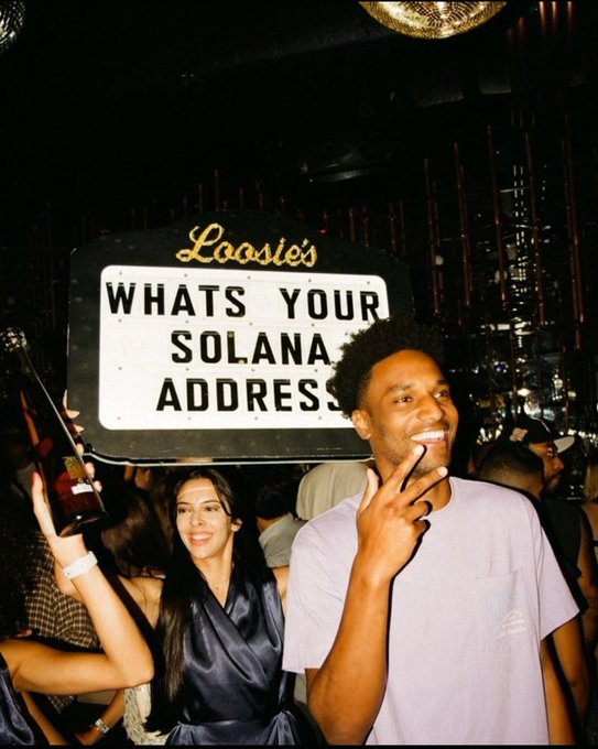 A young man in a white t-shirt smiles while holding up a sign that reads 'Loosie's WHATS YOUR SOLANA ADDRESS?' at a nighttime event.