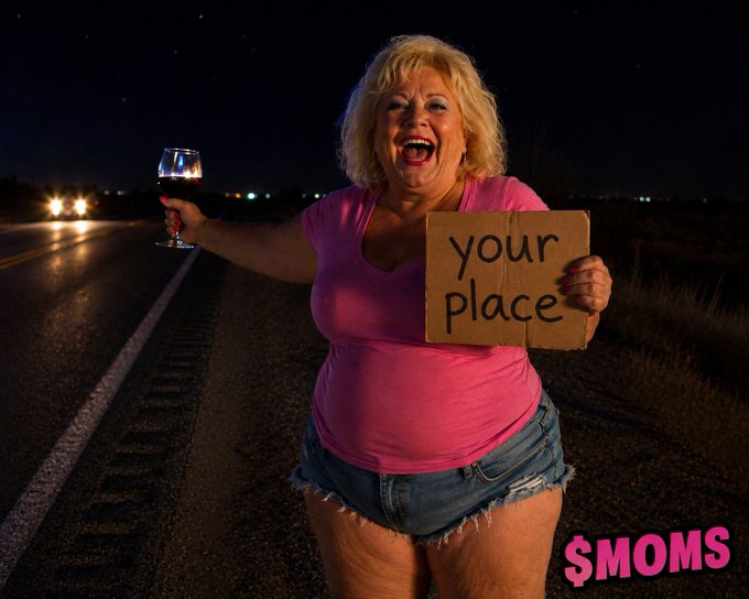 An older blonde woman in a pink shirt and denim shorts stands on a dark roadside holding a wine glass and a cardboard sign reading 'your place', with '$MOMS' text in the corner.