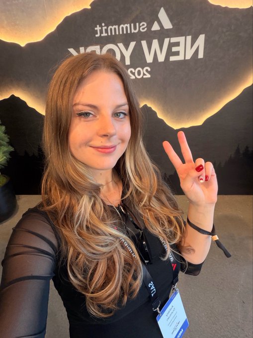 A selfie of a smiling woman with long blonde hair making a peace sign in front of a Summit 2025 New York backdrop, wearing a lanyard badge.