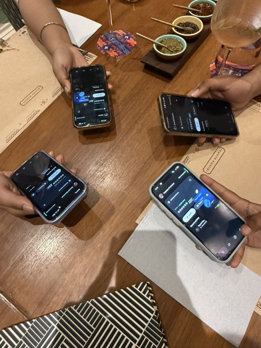 Four people sit around a wooden restaurant table, each holding a smartphone displaying chat or messaging app interfaces, with small condiment bowls and chopsticks visible in the background.