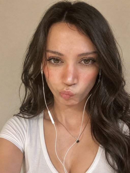 A selfie-style photo of a woman with long dark hair wearing earphones, a necklace, and a white t-shirt, making a kissing face at the camera.