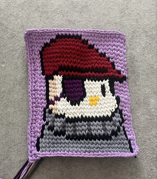 A small handcrafted plastic canvas or cross-stitch piece depicting a pixel-art character with dark hair, a red hat/cap, and a yellow detail, set against a purple background.