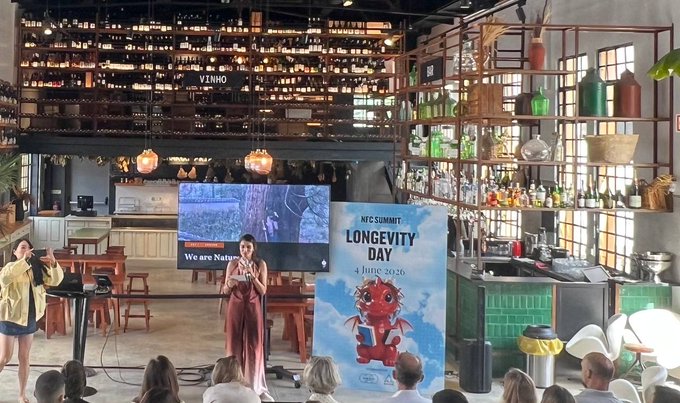 The image shows a woman standing at a podium with an audience gathered around her. She appears to be giving a speech or presentation, possibly about the "Longest Day" theme for the event. The room has a large screen displaying information related to the event, and there are several bottles placed on shelves in the background.