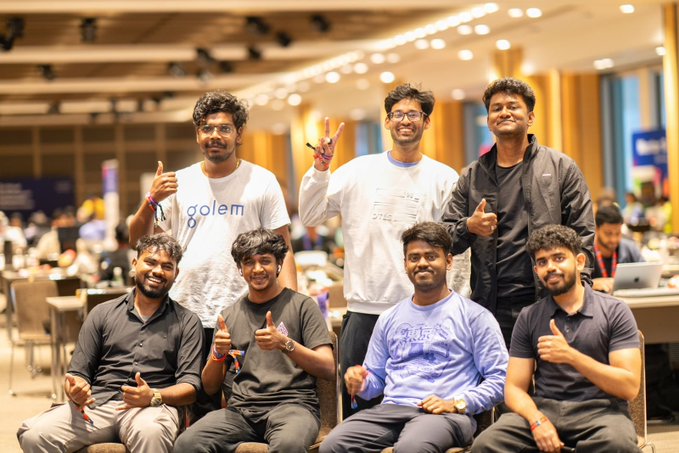 A group of six young men pose together at what appears to be a conference or event, with one wearing a shirt labeled 'golem' and others giving thumbs up.
