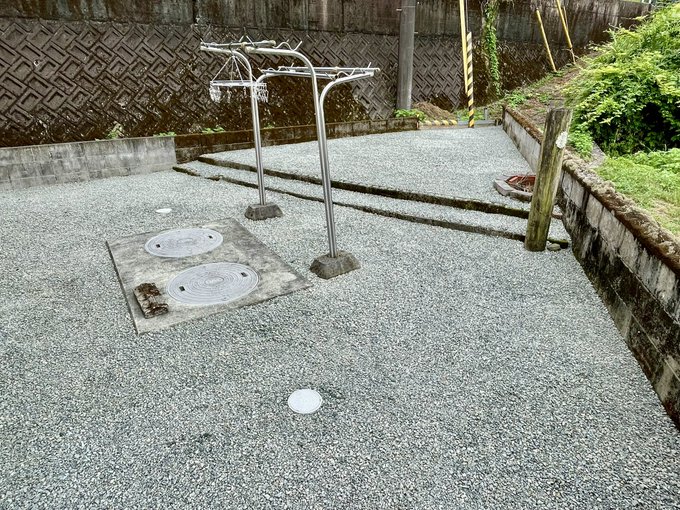 An outdoor public washing area features metal shower fixtures and two circular wash basins set into a concrete platform, surrounded by a retaining wall and greenery.