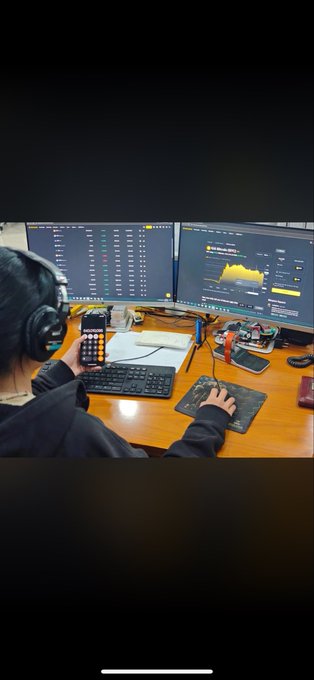 An over-the-shoulder view of a person wearing headphones at a desk with dual monitors showing financial trading charts and data, while their hand operates a mouse on a mousepad.