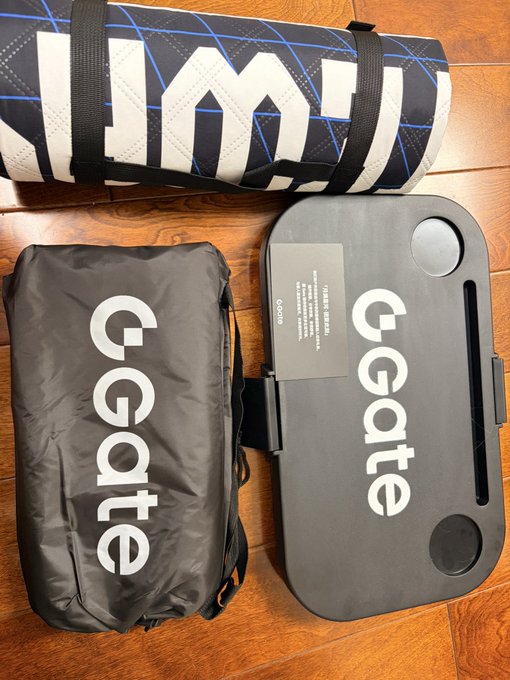 A camping kit from Gate.io featuring a black drawstring bag with the "Gate" logo and a foldable camping table with the Gate branding, along with a rolled-up mat or tent bag, all displayed on a wooden floor as part of a Mid-Autumn Festival gift package.