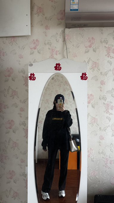 A person taking a mirror selfie while wearing a black tracksuit with 'DSQUARED2' written on the jacket, holding a phone, with red Chinese characters (福) decorating the top of the mirror.
