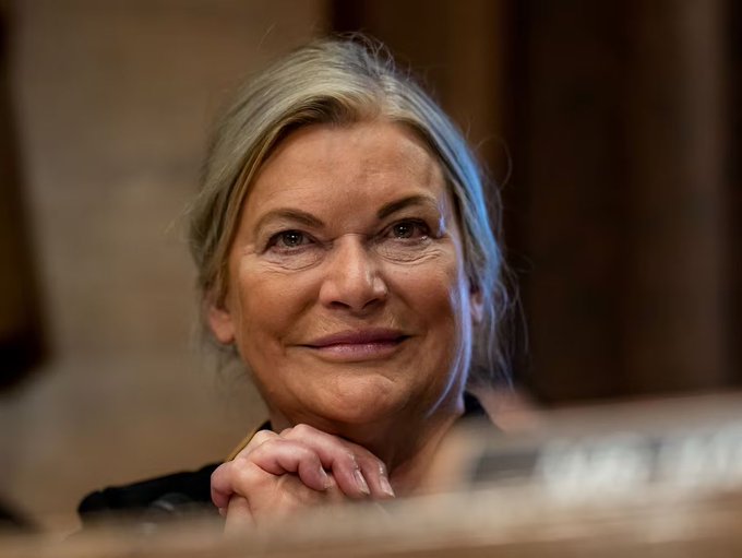 In the image, a woman with gray hair is sitting at a desk or table, smiling as she looks directly at the camera. The photo appears to be taken from behind her, giving an interesting perspective of her face. The caption below the image reads "BOOM! Sen. Lummis: “The CLARITY ACT passed committee." Trump is prepared to sign it into law," which suggests that this woman might have a connection with the political process or has been involved in some significant event related to the clarity act.