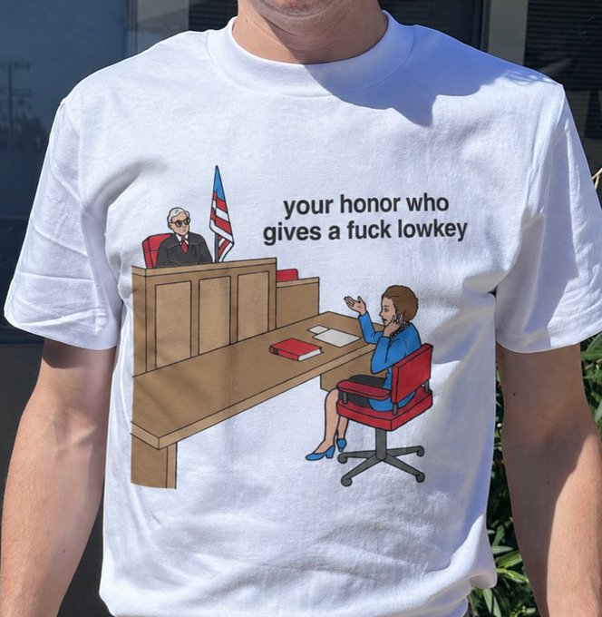 A white t-shirt features a cartoon graphic of a woman in court addressing a judge, with the text "your honor who gives a fuck lowkey" printed above.