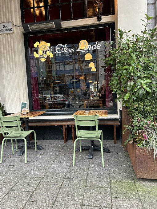 An outdoor seating area of a café called 'Een Eik en Linde' with green chairs, wooden tables, and a bench alongside large potted plants on a paved sidewalk.