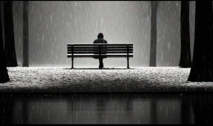 A black and white photograph of a person sitting alone on a park bench in falling rain, viewed from behind with reflections in water in the foreground.