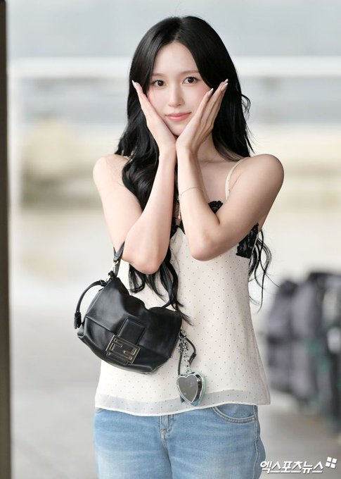 Korean celebrity (per '엑스포츠뉴스' watermark) posing with her hands on her cheeks, wearing a polka-dot cami top, jeans, and carrying a black handbag with a heart charm.