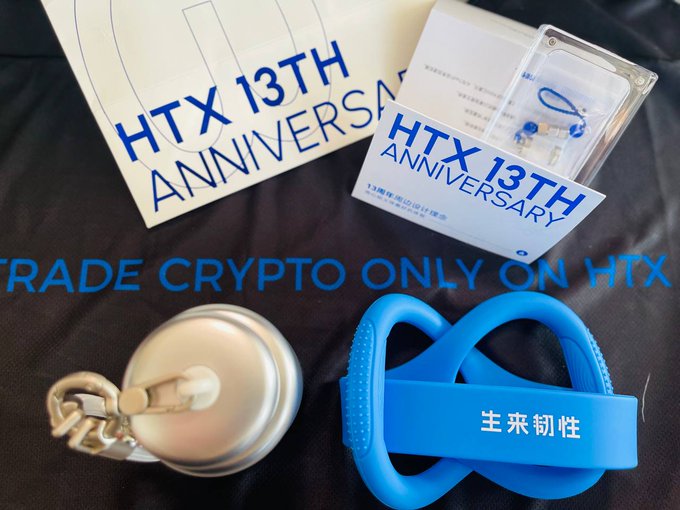 A promotional flat lay showcases HTX's 13th Anniversary gift box contents, including a silver portable cup, a twisted blue resistance band labeled "主来韧性," a "HTX 13TH ANNIVERSARY" card, and a commemorative gift bag, all arranged on a branded "TRADE CRYPTO ONLY ON HTX" mat. The image visually confirms the thoughtful array of fitness and lifestyle items mentioned in the review of Huobi's milestone celebration package.