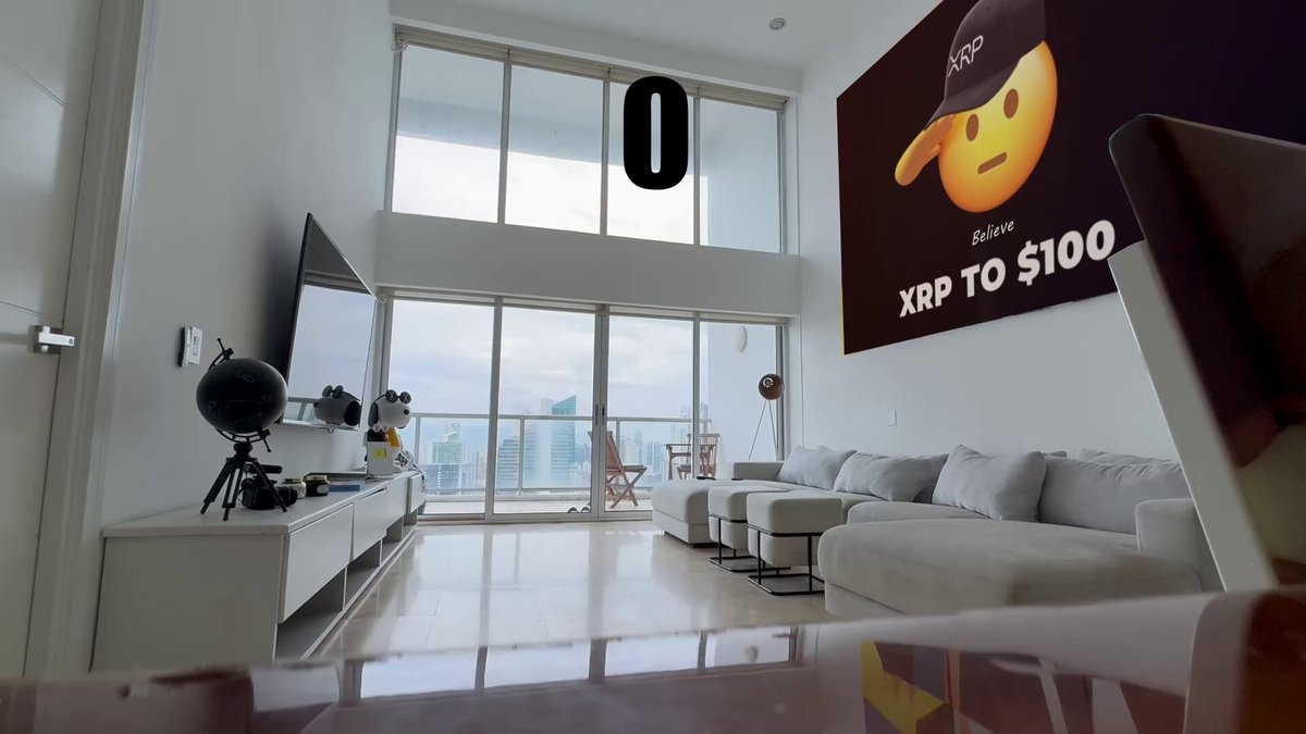 The image shows a large, open living room with white walls and furniture. There are two couches placed against the wall, one of which has an advertisement for Bitcoin on its back. A TV is mounted on the wall above the couch. The room also features a dining table in front of the couch and another smaller dining table nearby. On top of the larger dining table, there is a laptop open with a screen displaying the Bitcoin advertisement.