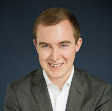 A professional headshot of a smiling young man with short brown hair wearing a grey suit jacket and white shirt against a dark blue background.