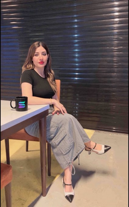 A woman sits at a white table in a black top and denim cargo pants next to a black mug with the Solana logo.