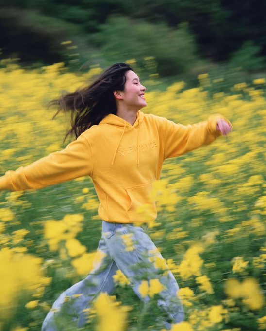A joyful woman in a yellow hoodie with "X EXI LOVE" text and light blue jeans runs freely through a vibrant field of yellow flowers, arms outstretched and smiling as she embraces the springtime beauty. Her flowing hair and radiant expression perfectly capture the carefree, exuberant spirit of celebrating "el día de la primavera."