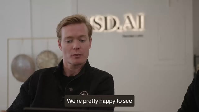 In this image, a man wearing a black jacket and a ponytail is seated at a desk with a laptop in front of him, facing away from the camera. Behind the man, there's an office setting with several other people visible in the background. The text on the image reads "We're pretty happy to see your interests". This suggests that the image may be related to a positive experience or satisfaction in exploring one's personal interests or hobbies through online platforms like the ones represented by the words seen in the post.