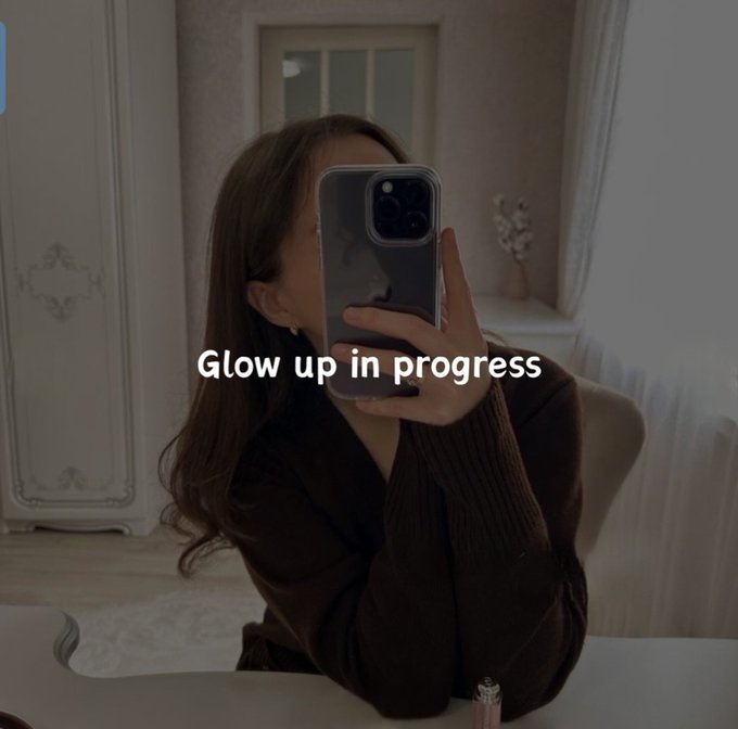 A mirror selfie of a woman with long brown hair wearing a dark brown sweater, holding up an iPhone that covers her face, with the text "Glow up in progress" overlaid on the image.