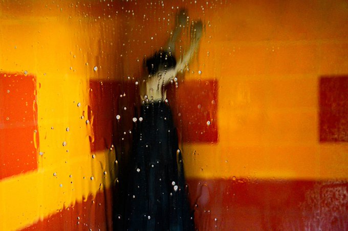 A blurred, atmospheric photograph captures the silhouette of a figure with raised arms seen through a rain-streaked glass pane, set against a backdrop of warm orange and red geometric light.