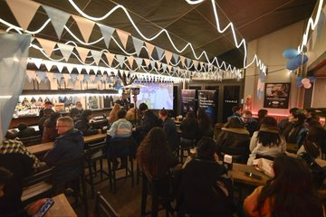 A dimly lit bar or restaurant interior decorated with string lights and bunting flags, with patrons seated at the bar and tables watching something on a screen.