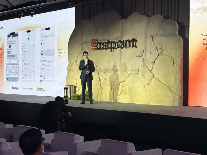 A man in a suit speaks on a microphone at an Eastpoint conference, with a stage backdrop resembling a large rock displaying the 'Eastpoint' logo and screens showing Korean mobile app interfaces and text 'AI가 대답을' (AI answers).