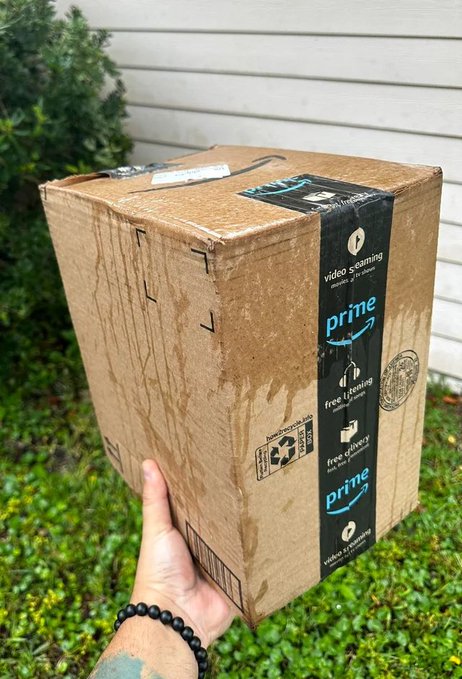 A person's hand holds a wet, rain-soaked Amazon Prime cardboard delivery box outdoors near a house with white siding.